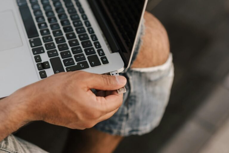 Hands inserting a USB drive into a laptop on the knees of a person wearing shorts, outdoors.